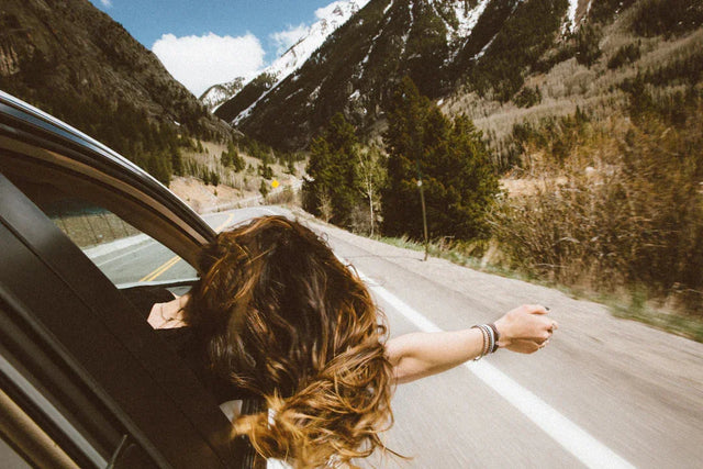 Person with long wavy brown hair leaning out of a car window on scenic mountain road
