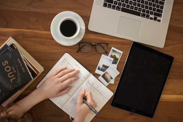 Hands writing a compelling school vision beside a laptop, coffee, and tablet.