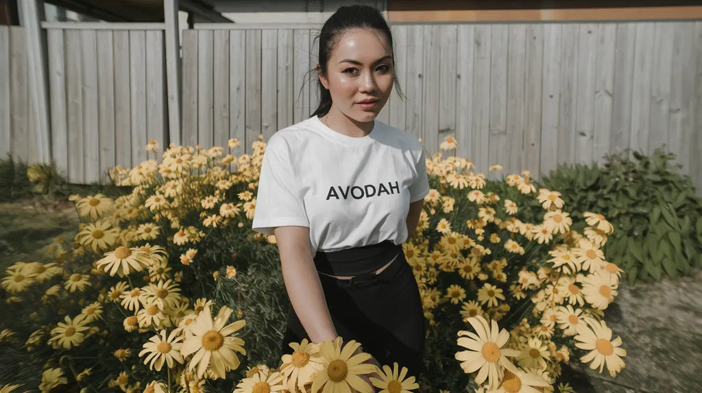 Person in a white AVOCADO t-shirt and black pants surrounded by yellow daisies, embracing limitless living.