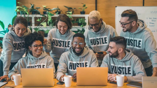 Group of coworkers wearing matching ’Hustle’ sweatshirts while looking at laptops together.